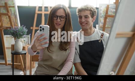 Die Frau hält das Smartphone für Selfie mit dem Maler, der eine Schürze trägt und im Atelier neben Leinwand und Staffelei sitzt, lächelt das Gerät an; geteilte Freude. Stockfoto