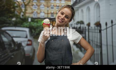 Verkäuferin in Denim-Schürze, die Eisbecher mit Handschuhen auf der Straße hält, lächelt den Betrachter an; fröhlicher Service. Stockfoto