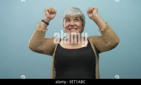 Frau, die Arme hebt und lächelt, mit freiliegenden Schultern und Creolen vor blauem Studiohintergrund, mit Strickjacke und Tanktop; Freude feiern Stockfoto