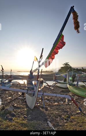 Traditionelle hölzerne Auslegerboote, die an einer felsigen Küste mit Blick auf einen hellen Sonnenuntergang über dem Meer in Bali liegen. Stockfoto