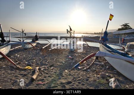 Traditionelle hölzerne Auslegerboote, die an einer felsigen Küste mit Blick auf einen hellen Sonnenuntergang über dem Meer in Bali liegen. Stockfoto
