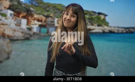 Junge Frau zeigt mit dem Zeigefinger auf die Kamera am Strand, trägt Brille und Denim, lange Haare sichtbar, lächelt sanft; verspieltes Selbstvertrauen. Stockfoto
