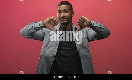Der junge schwarze Mann trägt ein graues Denim-Hemd und ein schwarzes T-Shirt und zeigt die Daumen nach unten mit beiden Daumen im roten Studio-Hintergrund. Stockfoto
