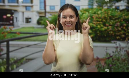 Die Frau zeigt beide Zeigefinger nach oben vor den Eingang des Gebäudes und lächelt in die Kamera; Optimismus wird ermutigt. Stockfoto
