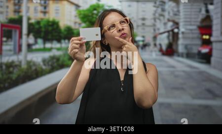 Frau mit weißer Kreditkarte zeigt Finger zum Kinn auf der geschäftigen Straße der Stadt; durchdachte Kaufentscheidung. Stockfoto