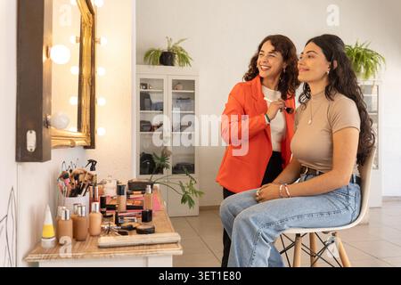 Make-up-Künstler und Kunde teilen sich einen freudigen Moment und lachen gemeinsam nach einer abgeschlossenen Make-up-Sitzung in einem hellen Schönheitssalon. Stockfoto