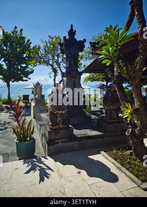 Traditionelle Steinstatuen und ein gestufter Schrein in einem Innenhof eines Seebades mit Blick auf das blaue Meer unter klarem Himmel in Sanur, Bali Stockfoto