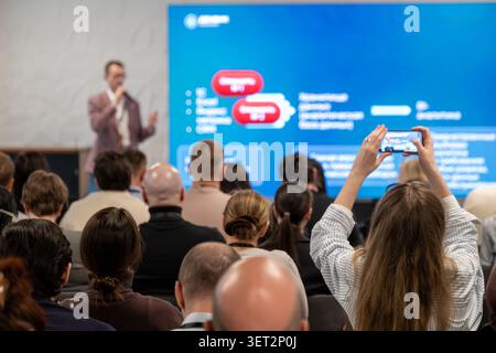 Das Konferenzpublikum beobachtet den Sprecher, der Teilnehmer nimmt die Präsentation am Telefon auf, professionelle Veranstaltungsatmosphäre. Stockfoto