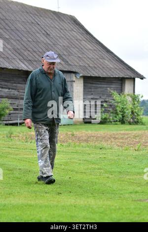 Ein älterer Bauer, der über ein ländliches Gehöft in der Nähe einer traditionellen Holzscheune spaziert. Lebensstil auf dem Land, landwirtschaftliches Erbe, Arbeit im Freien und friedliche Umgebung Stockfoto