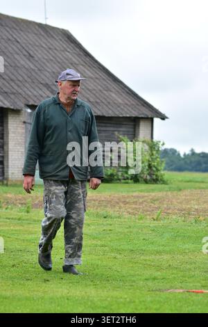 Ein älterer Bauer, der über ein ländliches Gehöft in der Nähe einer traditionellen Holzscheune spaziert. Lebensstil auf dem Land, landwirtschaftliches Erbe, Arbeit im Freien und friedliche Umgebung Stockfoto