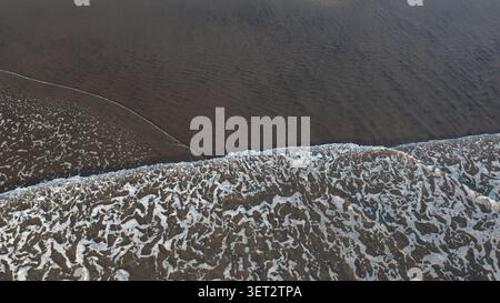 Blick aus der Vogelperspektive auf weißen Meeresschaum, der über dem dunklen vulkanischen schwarzen Sandstrand in Piha, Auckland, Neuseeland fließt. Stockfoto