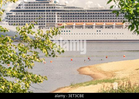Das Sommerleben am Flussufer mit entspannenden Menschen am Stadtstrand, während ein MSC-Kreuzfahrtschiff entlang der Daugava in Riga, Lettland, fährt. Stockfoto