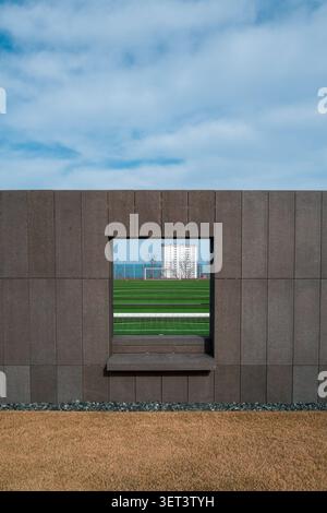 Ein symmetrischer Blick auf ein grünes Fußballfeld und das Meer wurde durch ein quadratisches Fenster in einer braunen Steinmauer perfekt eingerahmt. Stockfoto