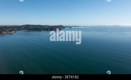 Aus der Vogelperspektive auf das ruhige blaue Wasser von Orewa Beach mit sanften grünen Hügeln und Küstenklippen unter klarem Himmel in Orewa, Auckland, Neuseeland. Stockfoto