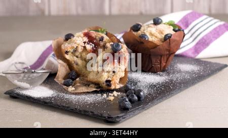 Zwei frisch gebackene Heidelbeermuffins werden auf einem dunklen Schieferbrett ausgestellt, einer davon wird geöffnet, um eine süße Marmelade freizulegen. Die Szene ist mit Puder übersät Stockfoto
