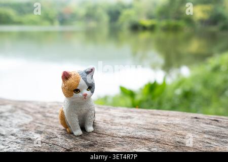 Chiang Mai, Thailand - 19. Juni 2026 : Eine Calico-Katzenfigur, die seitlich auf einem Holzstamm sitzt und schaut, mit einem See und grünem Wald im Hinterland Stockfoto