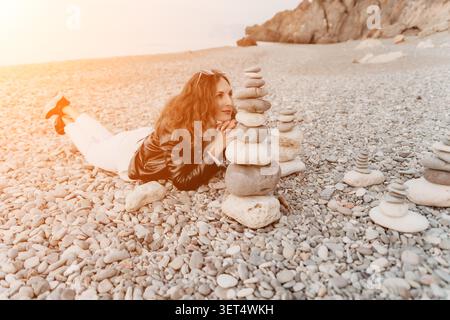 Frau Strandsteine entspannen an einem Kieselstrand in der Nähe von ausgewogenen cairns unter sanftem Sonnenlicht in einer Küstenlandschaft Stockfoto