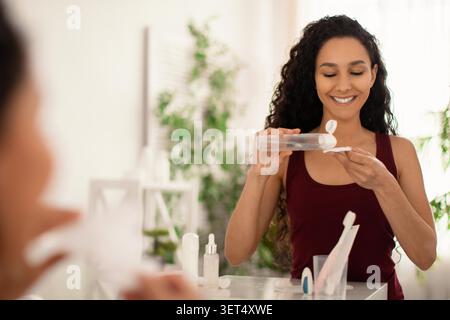 Happy Young Hispanic Woman Using Cosmetic Product At Badspiegel, Hautpflege Routine Stockfoto