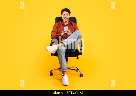 Lächelnder Junger Kaukasischer Mann, Der Im Bürostuhl Sitzt, Mit Smartphone, Gelber Hintergrund Stockfoto