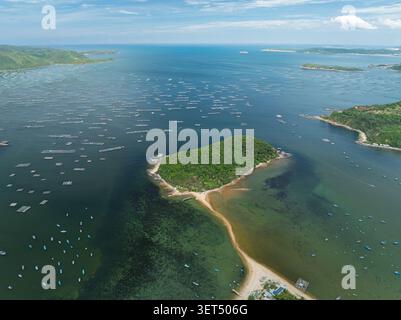 Luftpanorama der Küste mit einer kleinen bewaldeten Insel, Sandbank, zahlreichen Fischerbooten und Aquakulturkäfigen in klarem Flachwasser. Stockfoto