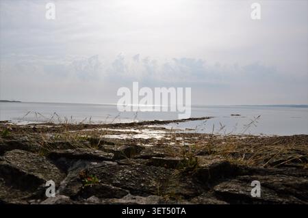Ein flacher Landschaftsblick auf ein ruhiges, schimmerndes Meer bei Ebbe hinter einer felsigen Steinmauer im Vordergrund unter einem bewölkten, hellen Himmel. Stockfoto