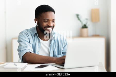 Der junge Mann arbeitet an einem Laptop in einem modernen Arbeitsbereich, lächelt mit angeschalteten Ohrhörern Stockfoto