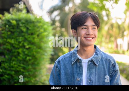 Horizontales mittleres Porträt eines fröhlich lächelnden jungen südostasiatischen Thai-Isan-Mannes in einer Jeansjacke, der mit grünem Bokeh nach draußen blickt Stockfoto