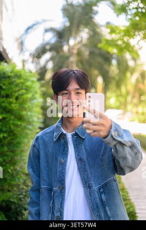 Vertikales mittleres Porträt eines fröhlich lächelnden jungen südostasiatischen Thai-Isan-Mannes in einer Jeansjacke, der sein Handy benutzt, um ein Selfie oder Vloggen in einem o zu machen Stockfoto
