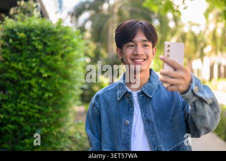 Horizontales mittleres Porträt eines fröhlich lächelnden jungen südostasiatischen Thai-Isan-Mannes in einer Jeansjacke, der sein Handy benutzt, um ein Selfie oder ein Vlogging-Outdo zu machen Stockfoto