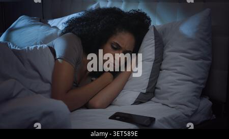 Schlafende afroamerikanische Frau wach mit lautem Handy Anruf wach im Bett in der Nacht ethnische Mädchen Frau wacht mit Smartphone Anruf Geräusche auf Stockfoto