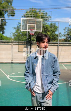 Vertikaler mittlerer Schuss eines nachdenklichen jungen südostasiatischen Thai-Isan-Mannes in einer Jeansjacke, der wegschaut, während er auf einem Basketballfeld posiert. Stockfoto
