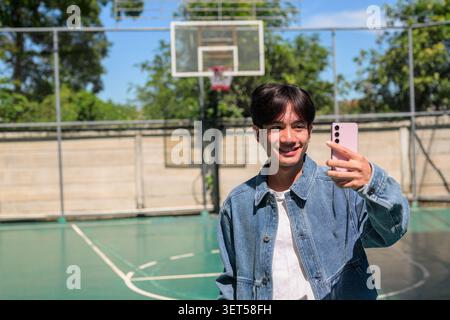 Horizontales mittleres Porträt eines fröhlich lächelnden jungen südostasiatischen Thai-Isan-Mannes, der mit seinem Handy ein Selfie auf einem Basketballfeld macht Stockfoto