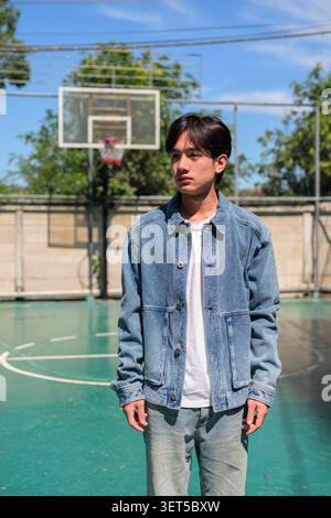 Vertikale mittlere Vollaufnahme eines ernsthaften nachdenklichen jungen südostasiatischen Thai-Isan-Mannes, der eine Jeansjacke und Jeans trägt und wegblickt, während er auf einem steht Stockfoto