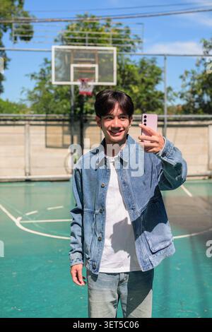 Vertikale mittlere Vollaufnahme eines glücklichen, lächelnden jungen südostasiatischen Thai-Isan-Mannes, der ein Selfie mit seinem Handy auf einem Basketballfeld macht. Stockfoto