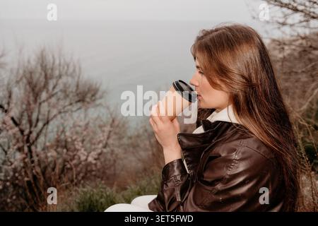 Kaffee Frau Küste junge Frau genießt ein warmes Getränk aus einem Pappbecher, während sie im Herbst am Meer sitzt Stockfoto