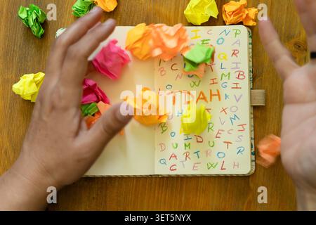 Hände werfen bunte zerknitterte Papierkugeln über ein offenes Notizbuch, gefüllt mit verstreuten Buchstaben und dem Akronym TDAH Stockfoto