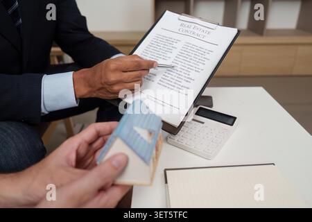 Ein Immobilienmakler erklärt einem Kunden einen Immobilienvertrag anhand eines Hausmodells. Konzept des Immobilienverkaufs und der professionellen Beratung. Stockfoto