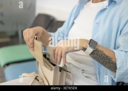Frau nimmt ihr weißes Smartphone vorsichtig aus einer Handtasche, zeigt ihre Smartwatch und ein detailliertes Flügeltattoo, was eine lässige und dennoch Technologie darstellt Stockfoto