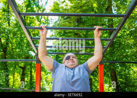 Mann mittleren Alters mit Augenklappe beim Pull-up an Affenbars im Outdoor-Park-Fitnessbereich Stockfoto