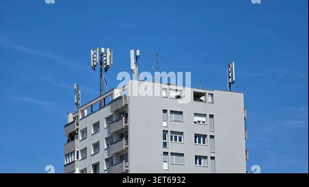 Sektorantennen und Funksender für Mobilfunknetze sind auf Metallhalterungen über dem Flachdach eines grauen Wohnblocks darunter montiert Stockfoto