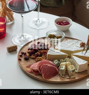 Holzbrett mit verschiedenen köstlichen Käsesorten, geschnittenem geräucherten Schinken, Oliven, Walnüssen, getrockneten Preiselbeeren, und Obstmarmelade, serviert mit einem Glas Rot Stockfoto