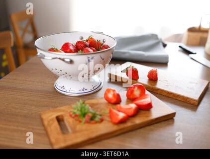 Schneiden Sie frische Bio-Erdbeeren auf Schneidebrettern aus Olivenholz in der Küche. Vintage-Sieb aus weißem Emaille gefüllt mit roten Sommerbeeren reflektierend Stockfoto