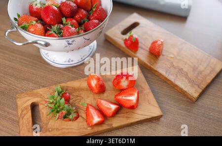 Schneiden Sie frische Bio-Erdbeeren auf Schneidebrettern aus Olivenholz in der Küche. Vintage-Sieb aus weißem Emaille gefüllt mit roten Sommerbeeren reflektierend Stockfoto