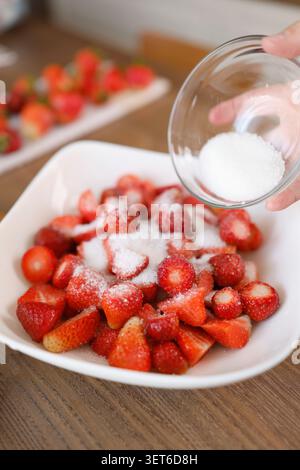 Weißen Zucker aus der Glasschale auf frisch geschnittene Erdbeeren in weißer Platte gießen. Traditionelle Routine zur Konservierung von Beeren im Sommer, die elegant ist Stockfoto