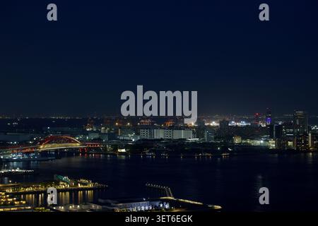 Kobe Hafen in der Abenddämmerung, mit der berühmten roten Kobe Brücke, die den Hafen überspannt, mit Gebäuden am Wasser, die vor dem Dämmerungshimmel beleuchtet sind. Stockfoto