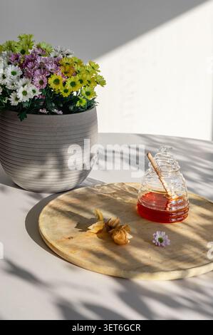 Nahaufnahme flacher Lagen eines hölzernen Honiglöffels mit Physalis, Gänseblümchen und einem Glas Honigglas auf einem sonnendurchfluteten Holzbrett. Ein natürliches, süßes Essen, mit dem Stillleben leben Stockfoto