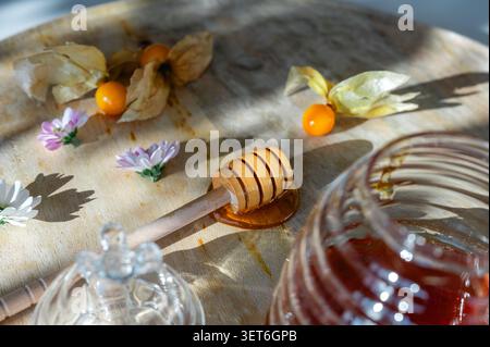 Nahaufnahme flacher Lagen eines hölzernen Honiglöffels mit Physalis, Gänseblümchen und einem Glas Honigglas auf einem sonnendurchfluteten Holzbrett. Ein natürliches, süßes Essen, mit dem Stillleben leben Stockfoto