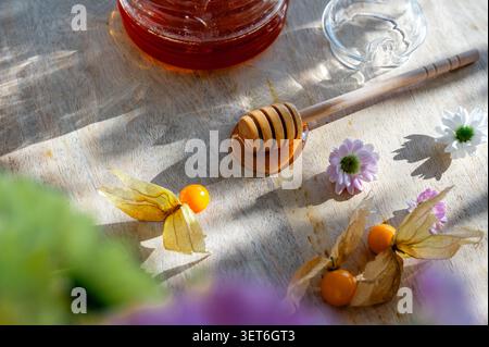 Nahaufnahme flacher Lagen eines hölzernen Honiglöffels mit Physalis, Gänseblümchen und einem Glas Honigglas auf einem sonnendurchfluteten Holzbrett. Ein natürliches, süßes Essen, mit dem Stillleben leben Stockfoto