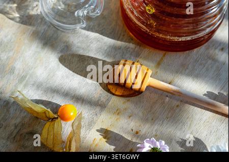 Nahaufnahme flacher Lagen eines hölzernen Honiglöffels mit Physalis, Gänseblümchen und einem Glas Honigglas auf einem sonnendurchfluteten Holzbrett. Ein natürliches, süßes Essen, mit dem Stillleben leben Stockfoto