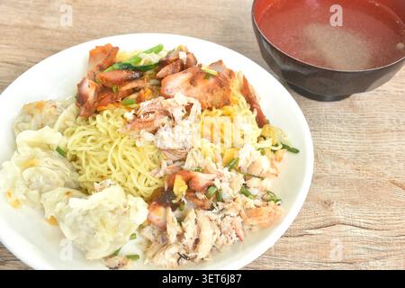 Trockene chinesische Ei gelbe Nudeln mit rotem Barbecue-Schweinefleisch und Knödel mit Krabbenfleisch auf Teller mit klarer Suppentasse Stockfoto